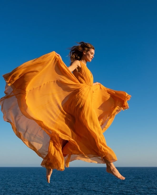 A dancer in a saffron gown leaping against a clear blue sky above the sea