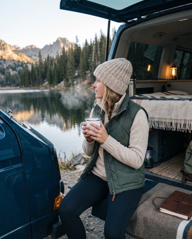 A woman with a coffee at an open tailgate, lake and mountains beyond
